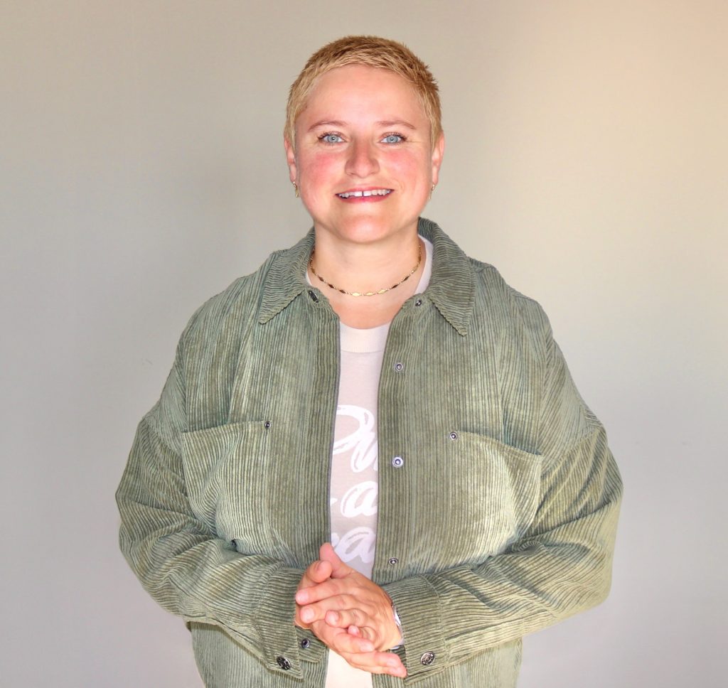 Coach Bea Schwaiger steht vor einer braunen Wand und lächelt in die Kamera. Sie hat beide Hände zusammengelegt.