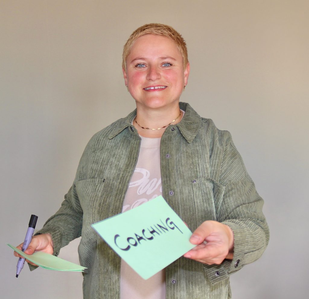 Themen im Coaching: Coach Bea Schwaiger lächelt in die Kamera. Sie hält einen Zettel in der Hand auf dem das Wort Coaching steht.