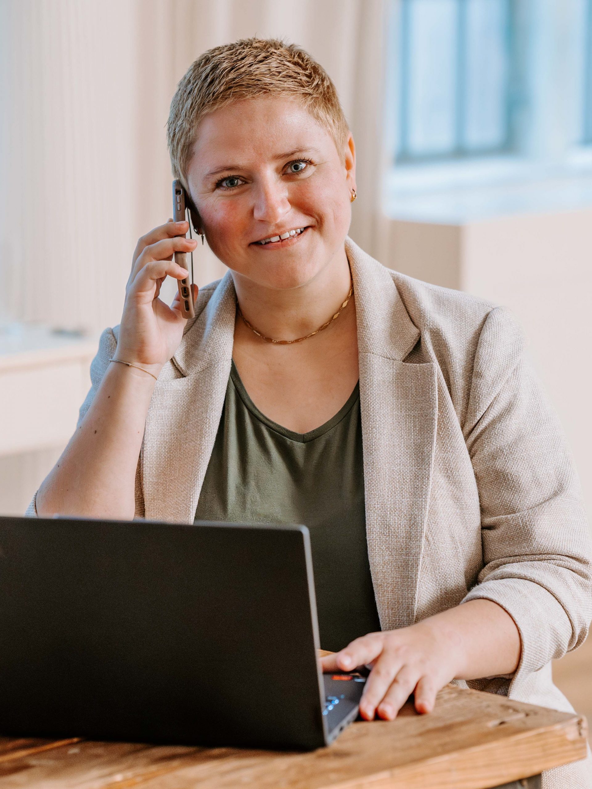 Coach Bea Schwaiger sieht lächelnd in die Kamera. Am rechten Ohr hält sie ihr Handy, vor sich auf dem Tisch hat sie ein aufgeklapptes Notebook stehen.
