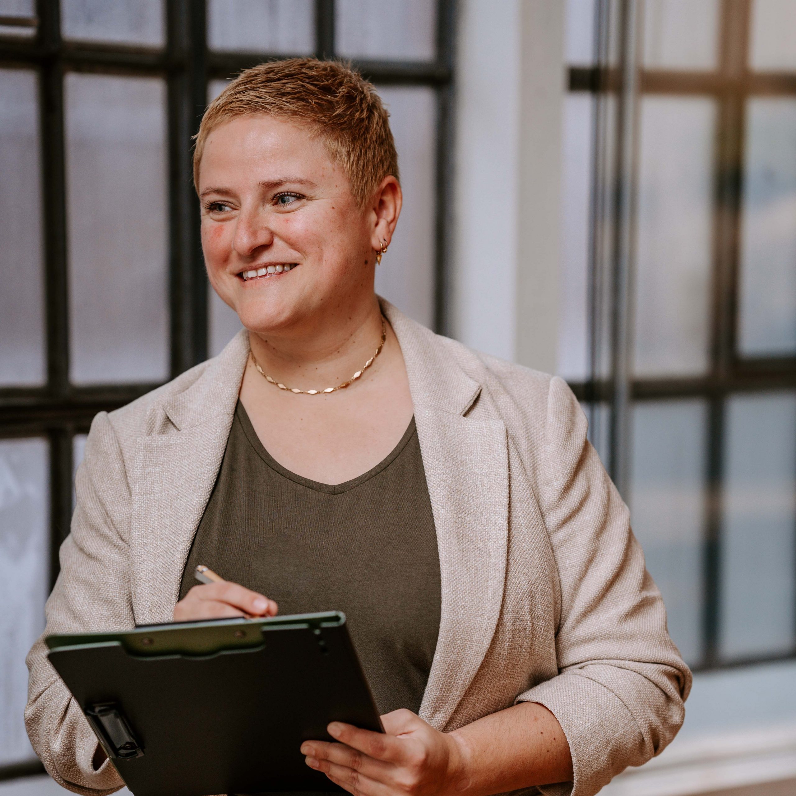 Coach Bea Schwaiger schaut zur Seite und lächelt. In der linken Hand hält sie ein Klemmbrett, in der rechten einen Stift und macht Notizen.